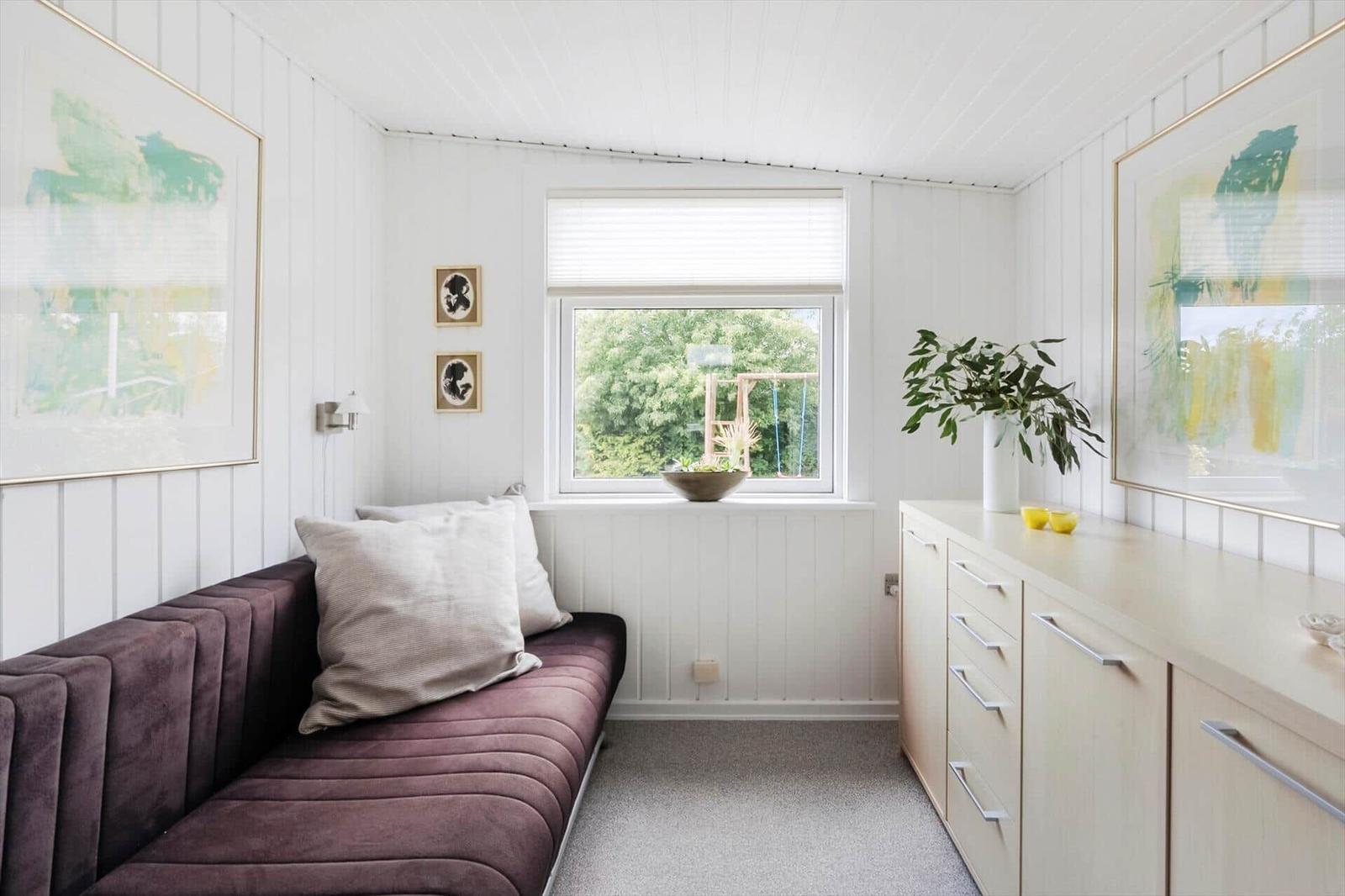 Ein Raum mit einem dunkelvioletten Sofa, weißen Wänden und einem Fenster mit Blick auf einen Garten.