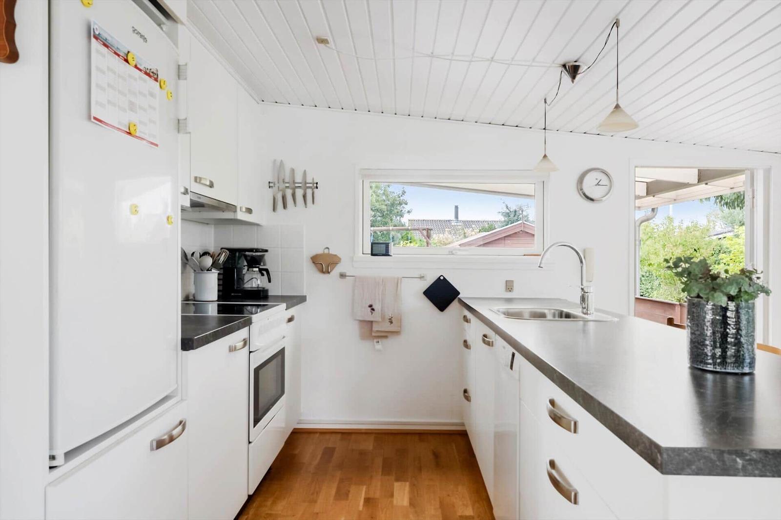 Küche mit weißen Schränken, dunkler Arbeitsplatte und Holzboden. Fenster und Tür zu Außenbereich.