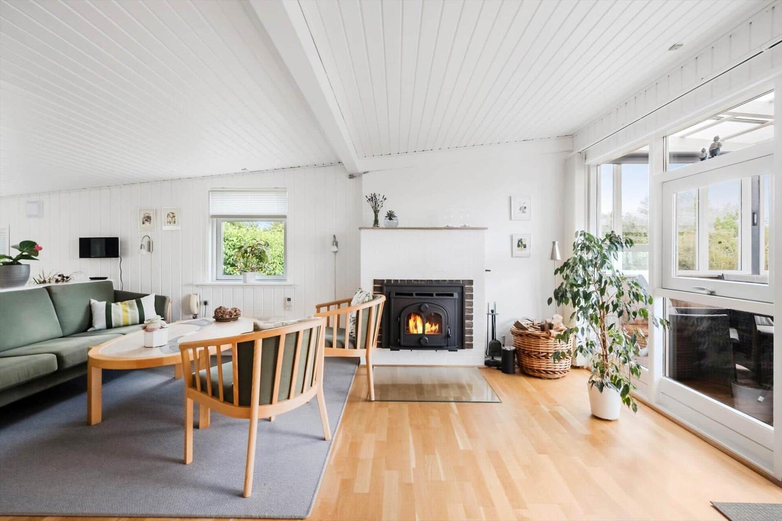 Wohnzimmer mit Kamin, Sofa, Stühlen und Holzboden. Große Fenster und Pflanzen.