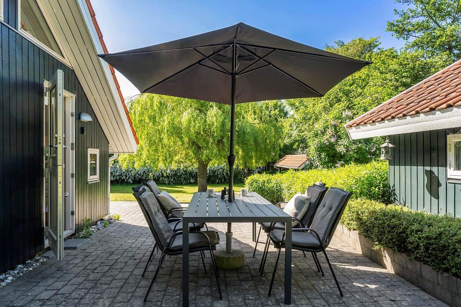 Terrace with table, chairs, and umbrella between houses and greenery.