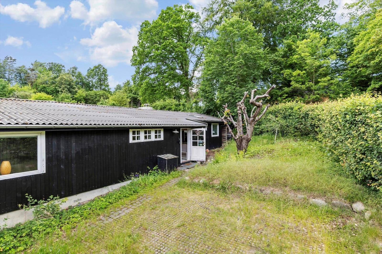 Black wooden house with white windows and door. Green lawn with trees and shrubs.