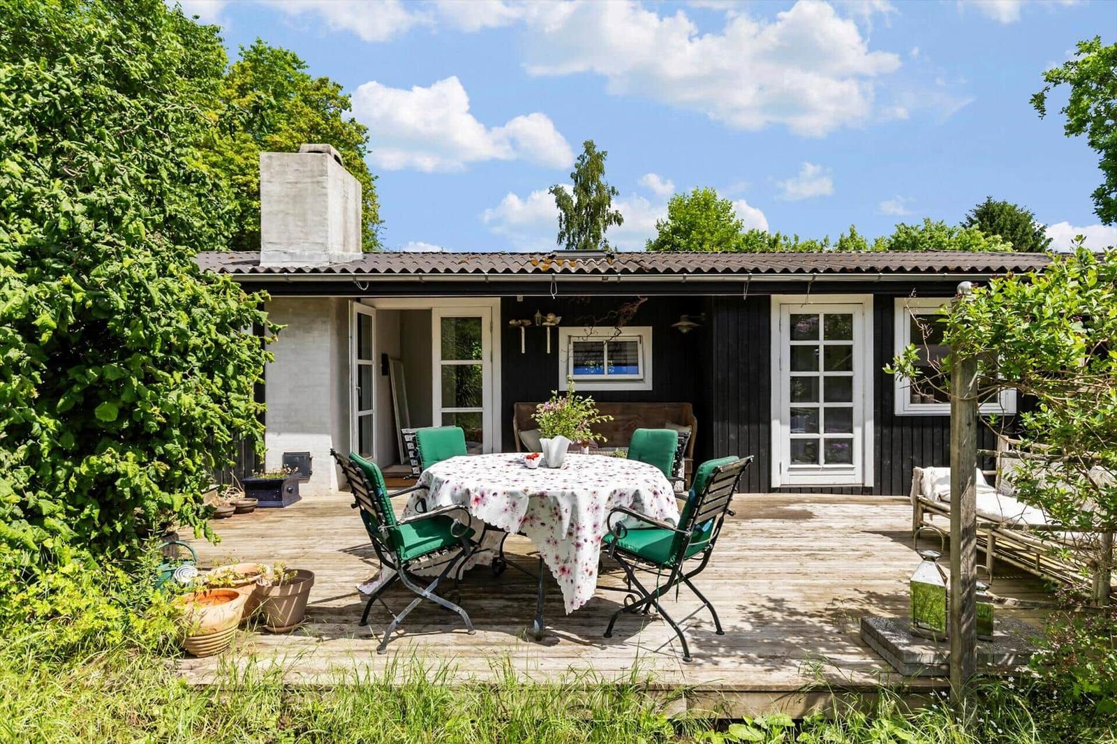 Deck with table, chairs, and plants in front of a house with dark wood and white windows.