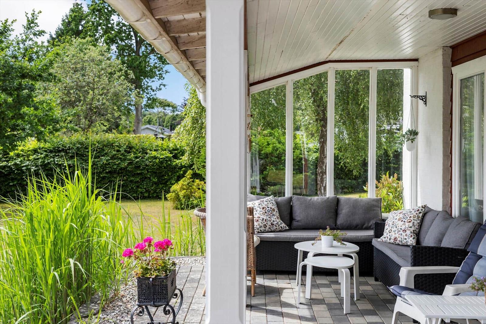 Patio with seating, glass walls, and garden view. White tables and plants.