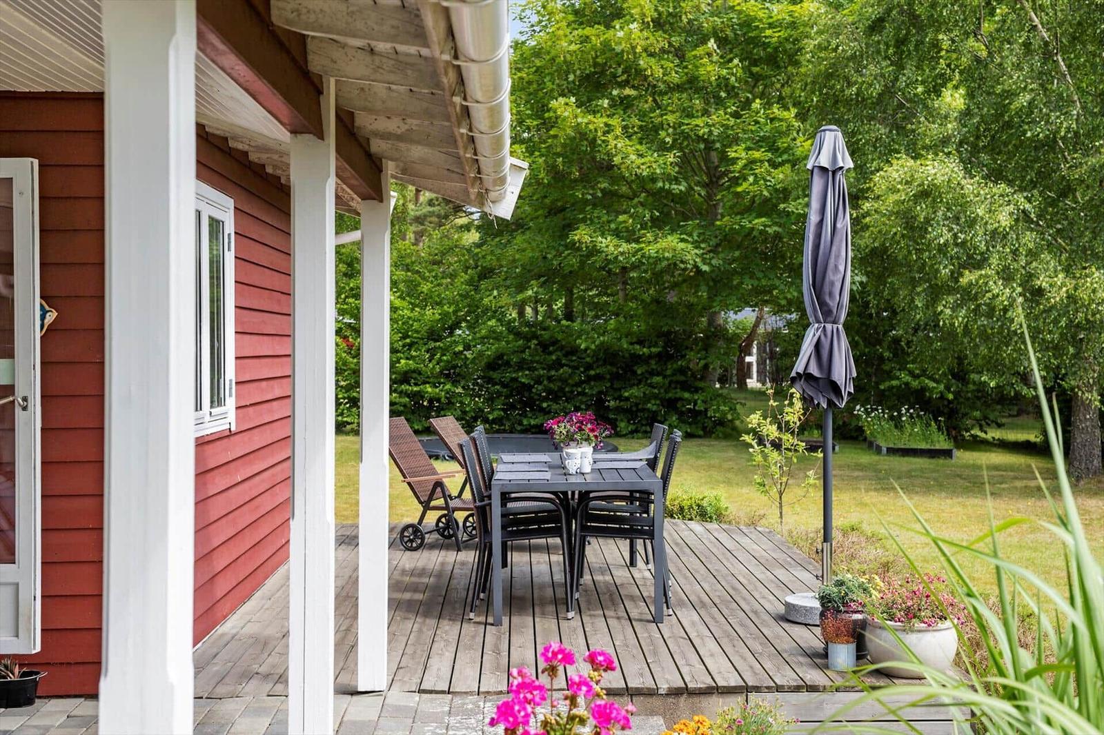 Deck with table, chairs, and umbrella beside red house with green garden.