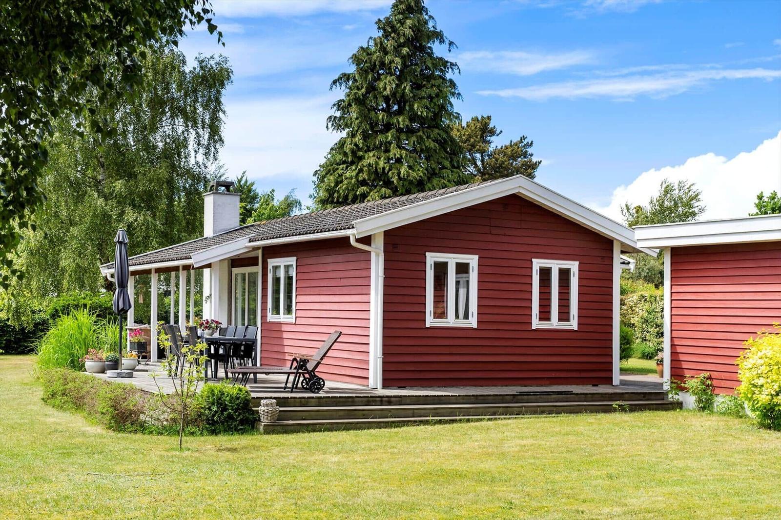Red wooden house with terrace, garden, and grill area under blue sky.