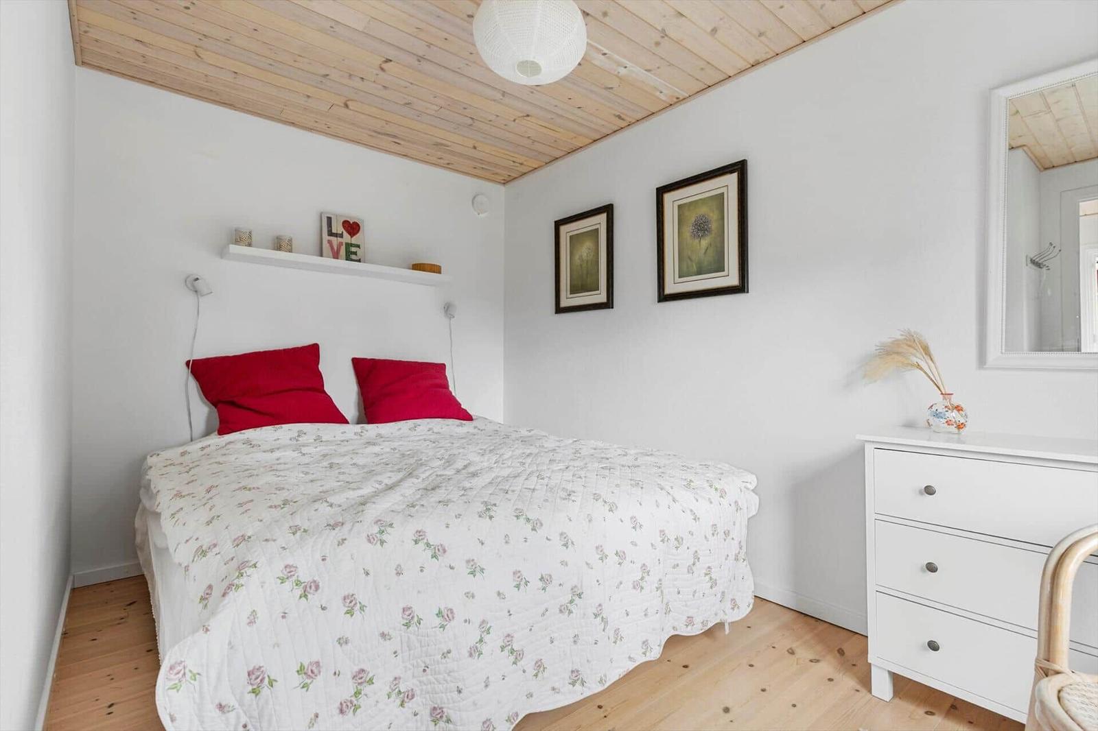 Bedroom with bed, white walls and wooden floor. Pictures and a mirror hang on the wall.