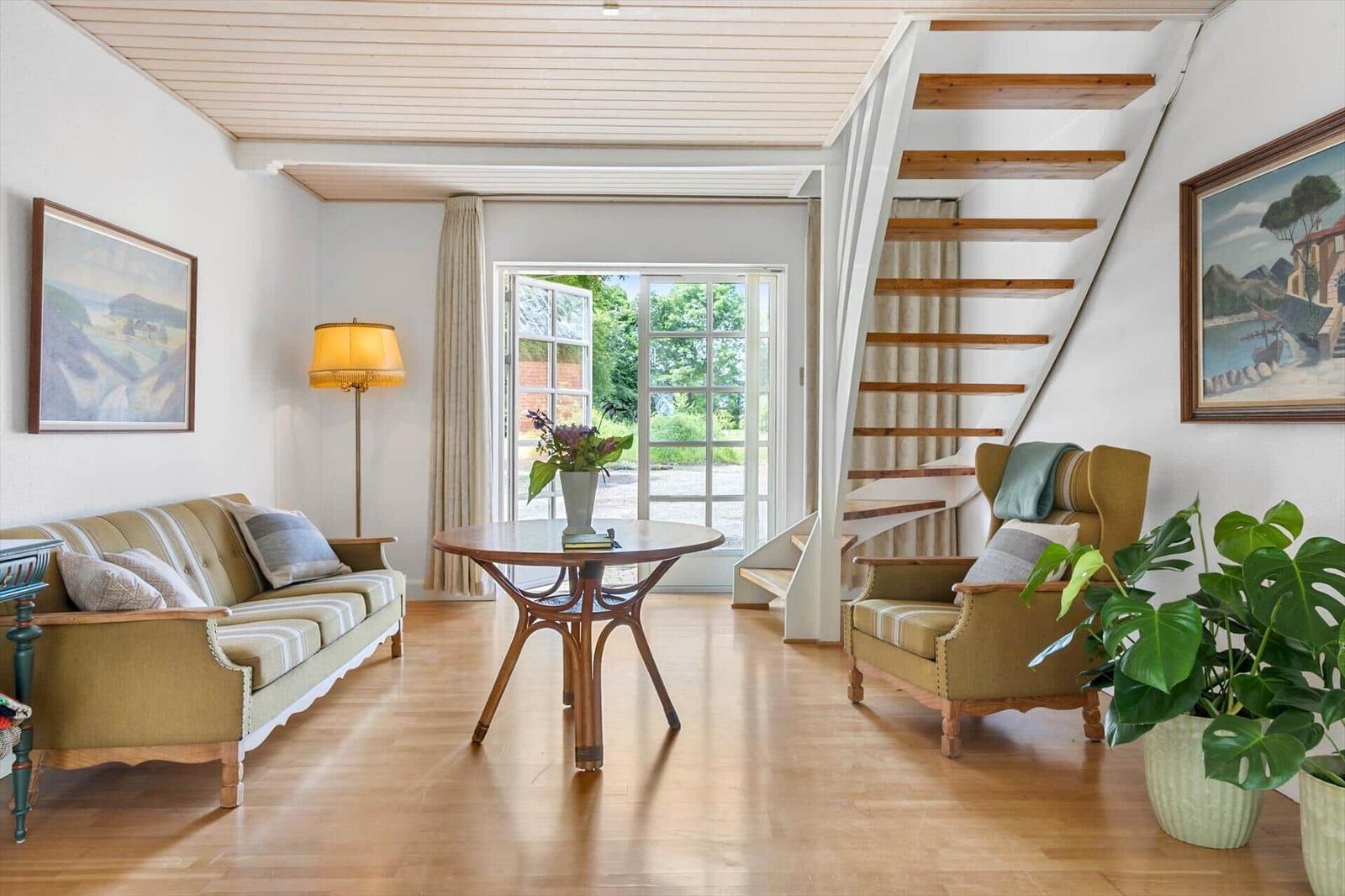 Wohnzimmer mit Sofa, Sessel, Tisch und Treppe. Fenster zur Außenseite.
