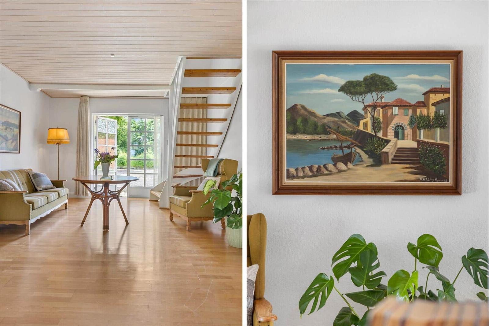 Wohnraum mit Holzboden, Sofa, Sessel und Treppe. Wandbild zeigt Küstenlandschaft.