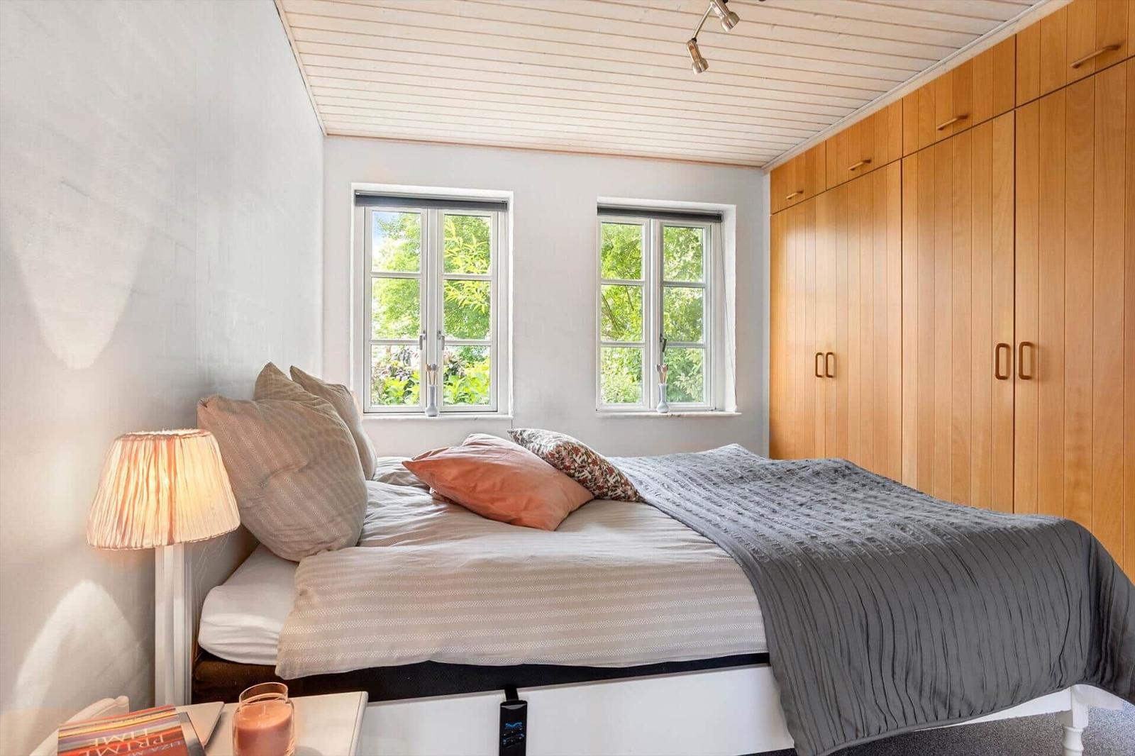 Schlafzimmer mit Bett, Nachttisch, Lampe und Holzschrank. Zwei Fenster zeigen grüne Bäume.