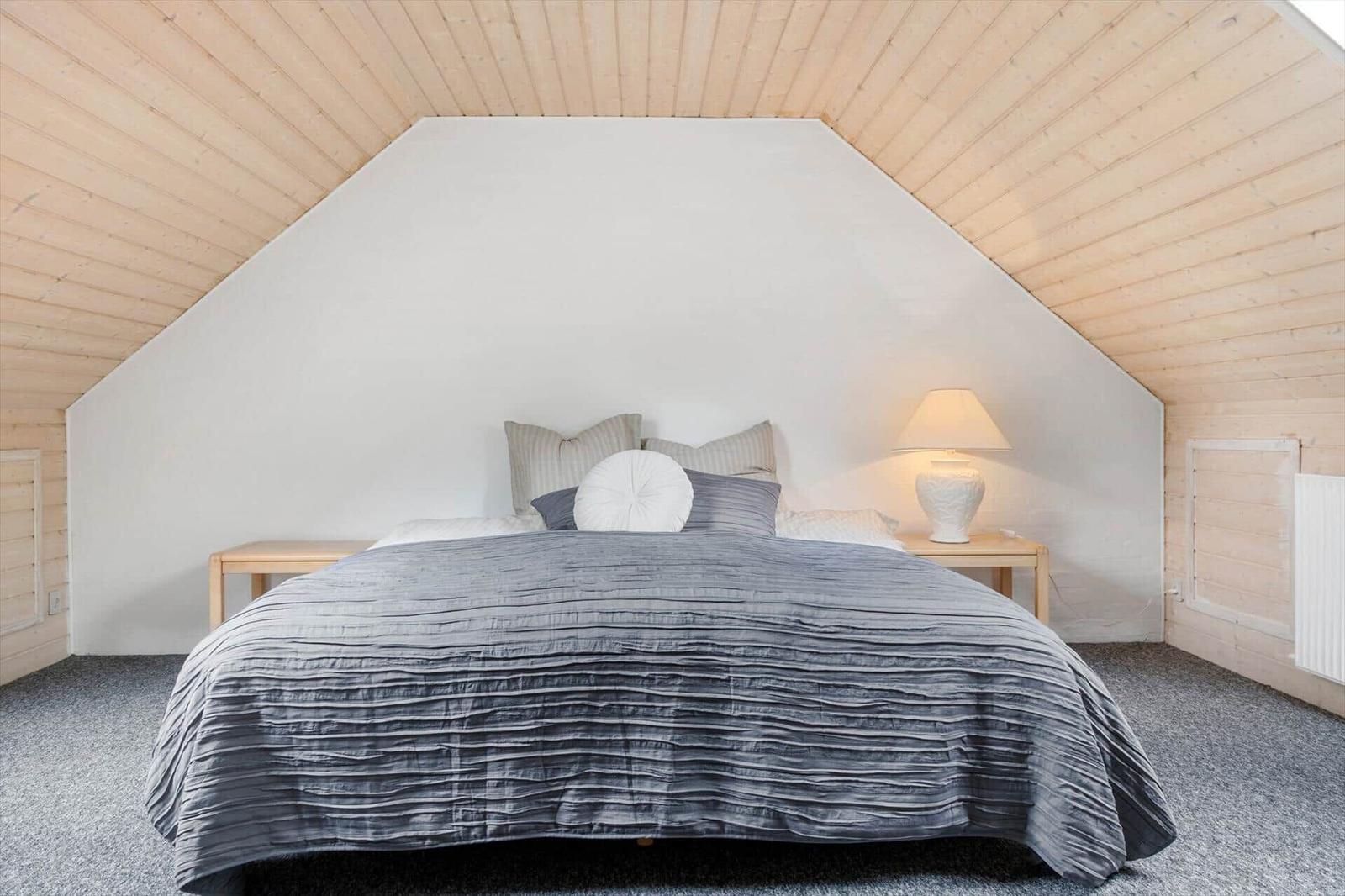 Schlafzimmer mit Dachschrägen, Bett mit grauer Bettwäsche und Nachttisch mit Lampe.