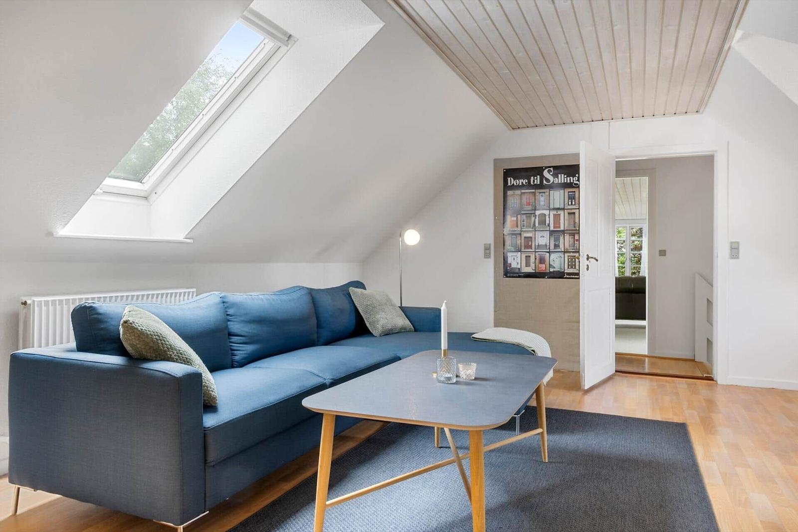 Ein Wohnzimmer mit blauem Sofa, Holztisch und Dachfenster. Holzdecke und Tür zum Flur.
