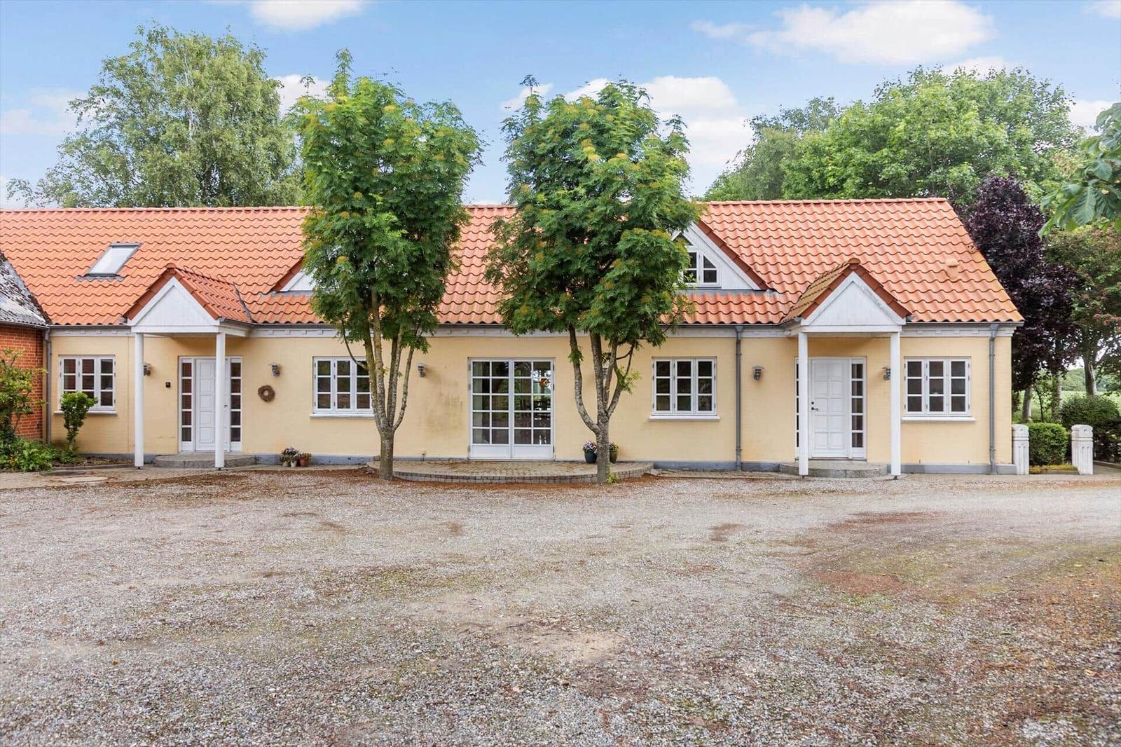 Ein gelbes Haus mit rotem Dach und mehreren Eingängen, umgeben von Bäumen und Kies.