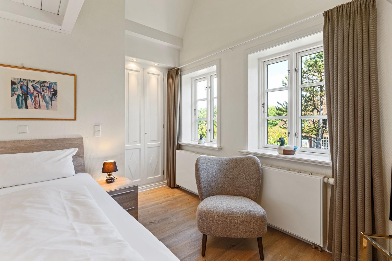 Schlafzimmer mit Bett, Stuhl und Fenstern. An der Wand hängt ein Bild.
