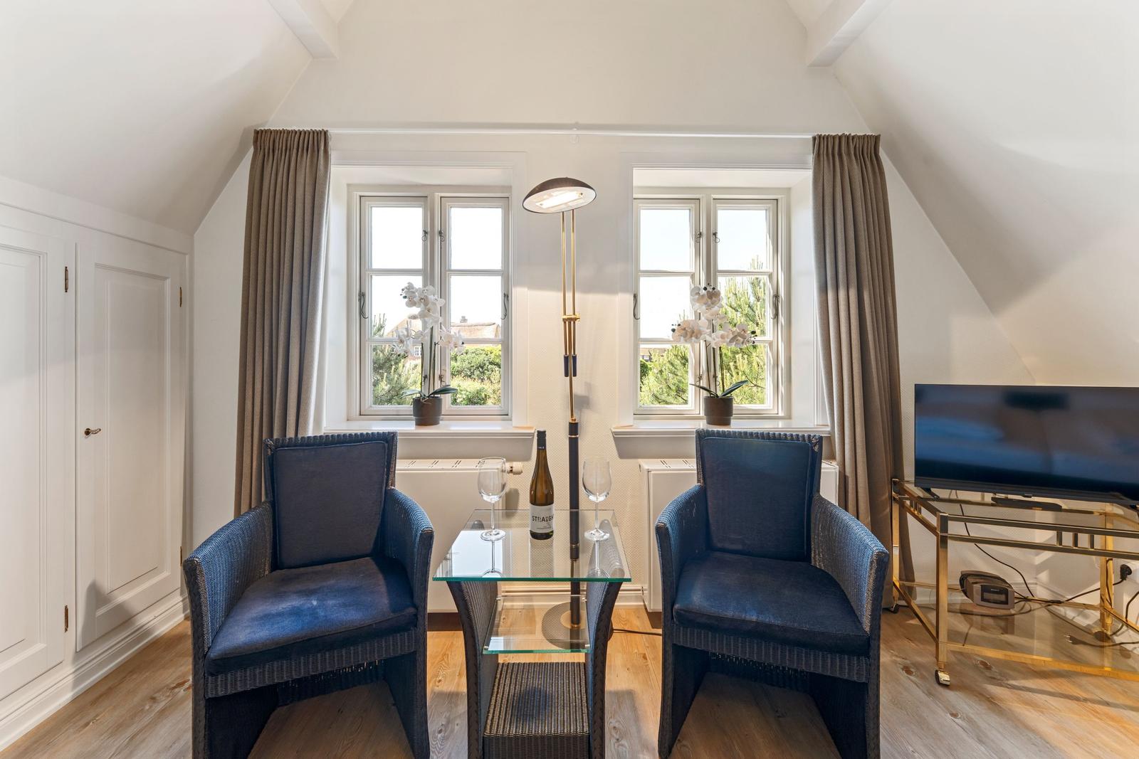 Zwei blaue Sessel stehen um einen Glas-Tisch. Fenster mit Vorhängen und Fernseher.