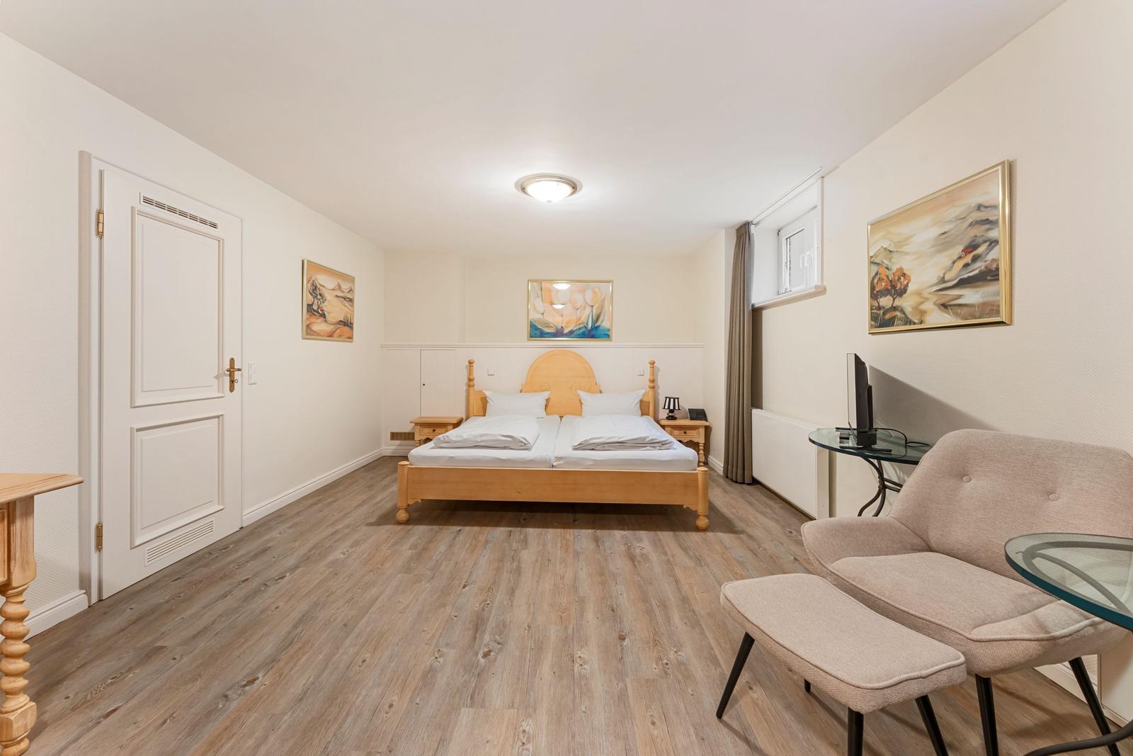 Schlafzimmer mit Doppelbett, Holzboden und Sitzgruppe. An der Wand hängen Bilder.
