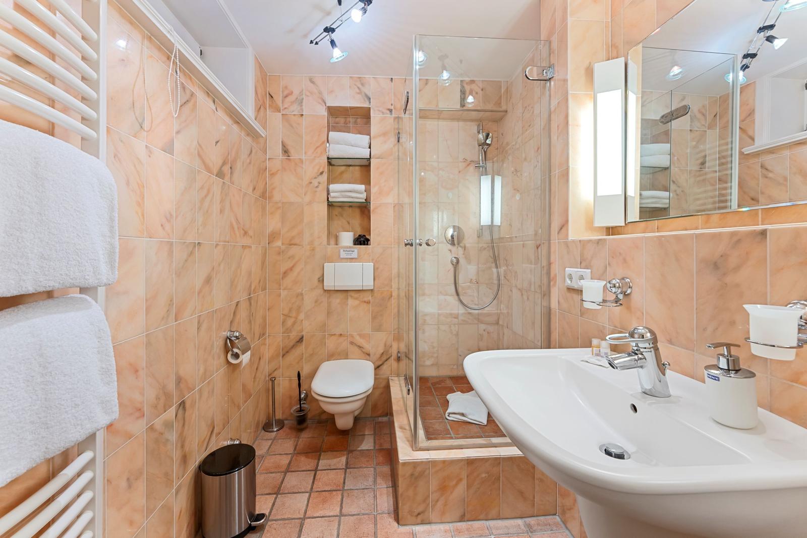 Badezimmer mit Dusche, Waschbecken und Toilette. Marmortile und weiße sanitäre Anlagen.