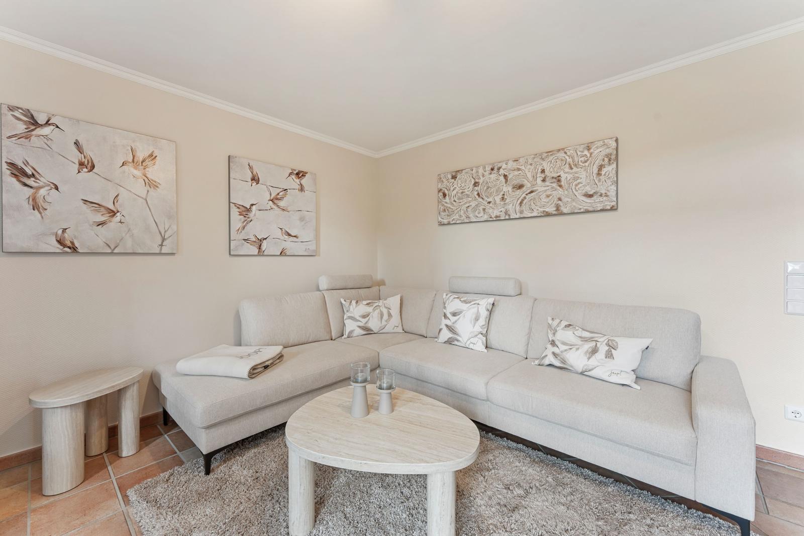Ein Wohnzimmer mit einem hellen Ecksofa, einem runden Tisch und Wandbildern.