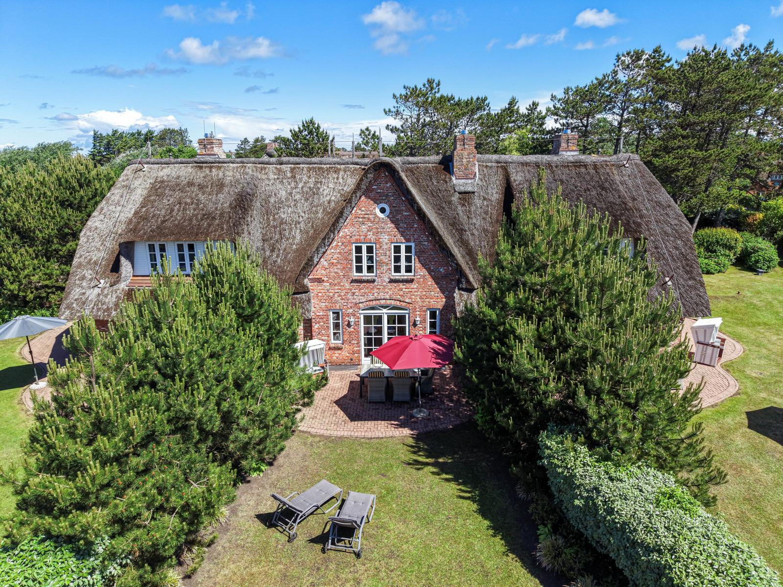 Haus mit Strohdach, Garten, Liegestühle und Terrasse mit Sonnenschirm.