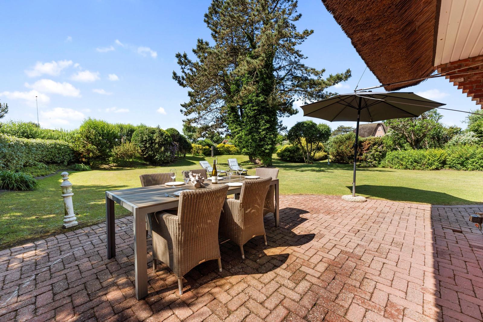 Terrasse mit Tisch, Stühlen und Sonnenschirm. Grünfläche und Baum im Hintergrund.