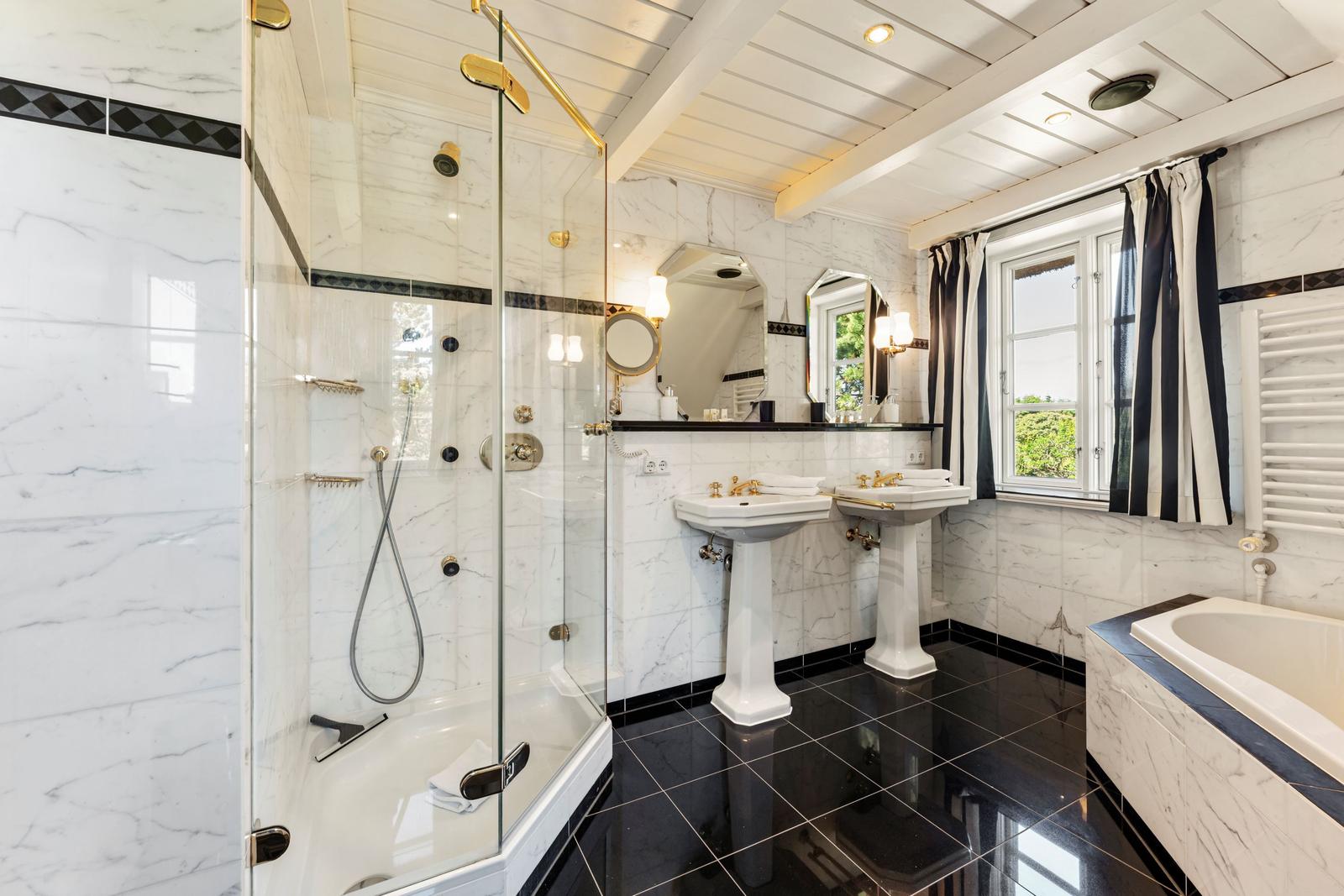 Badezimmer mit Dusche, zwei Waschbecken und Badewanne, weiße Marmorwände und schwarze Fliesen.