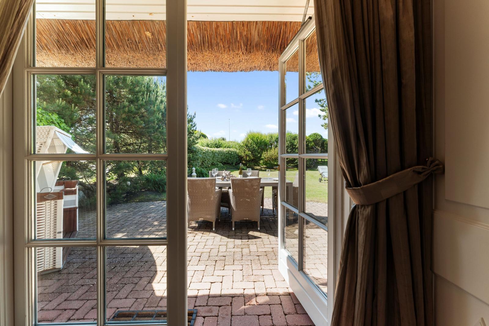 Durch die Glastüren blickt man auf eine Terrasse mit Tisch, Stühlen und einem Dach aus Stroh.