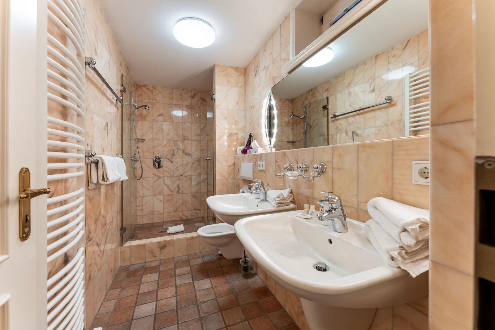 Badezimmer mit Dusche, zwei Waschbecken und Toilette. Wände mit Marmorfliesen verkleidet.