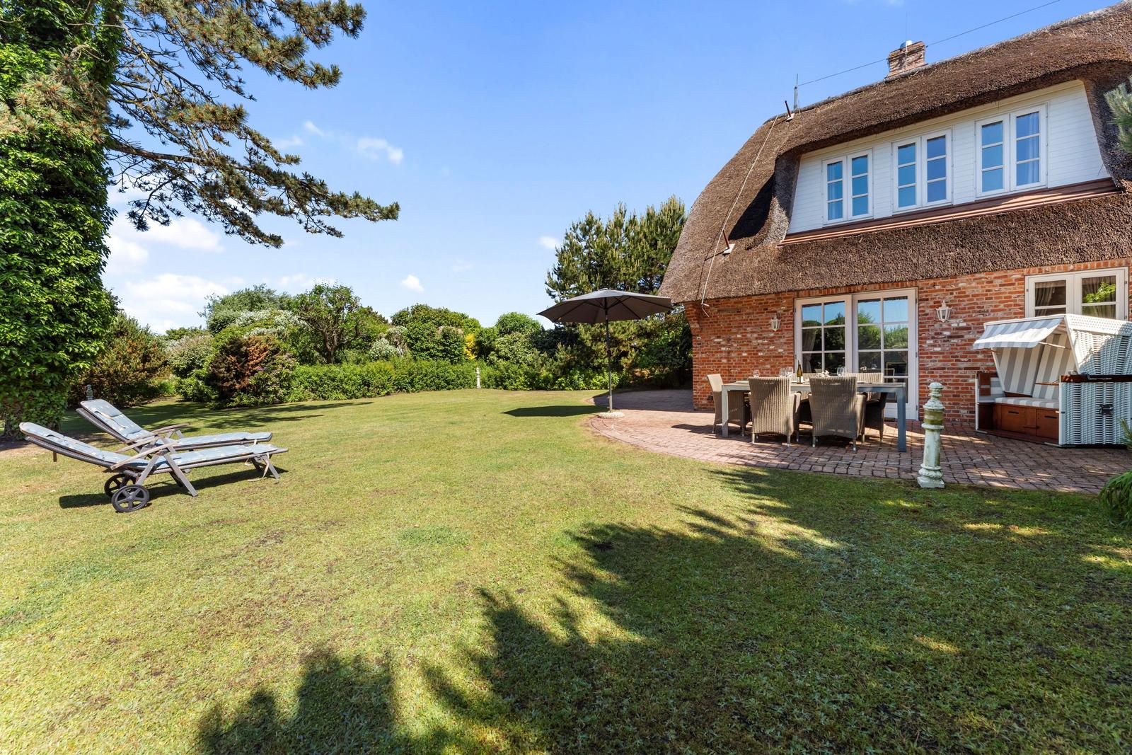 Haus mit strohgedecktem Dach, Garten mit Liegestühlen und Terrasse mit Tisch und Stühlen.