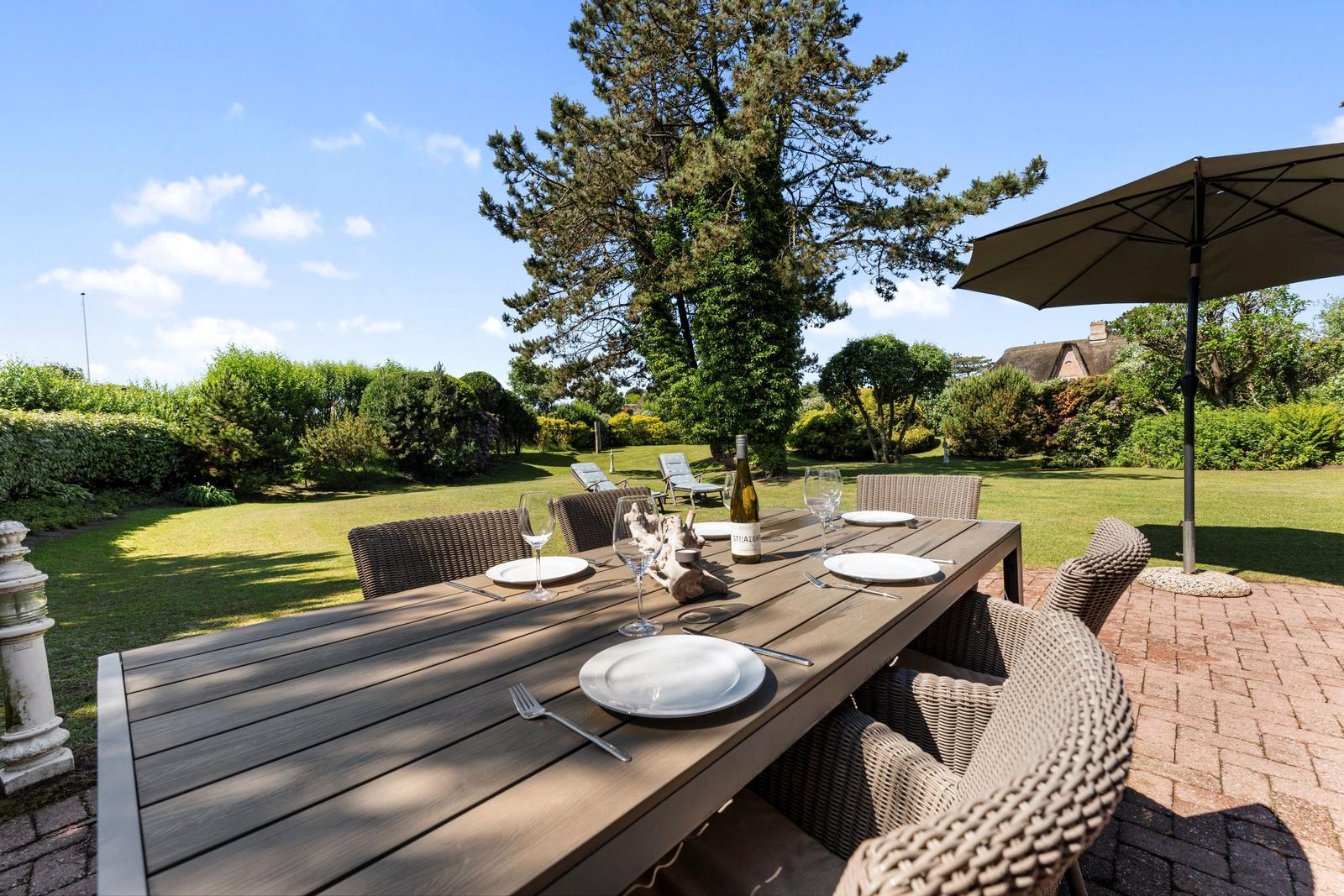 Garten mit Esstisch, Stühlen und Sonnenschirm. Grünflächen und Bäume im Hintergrund.