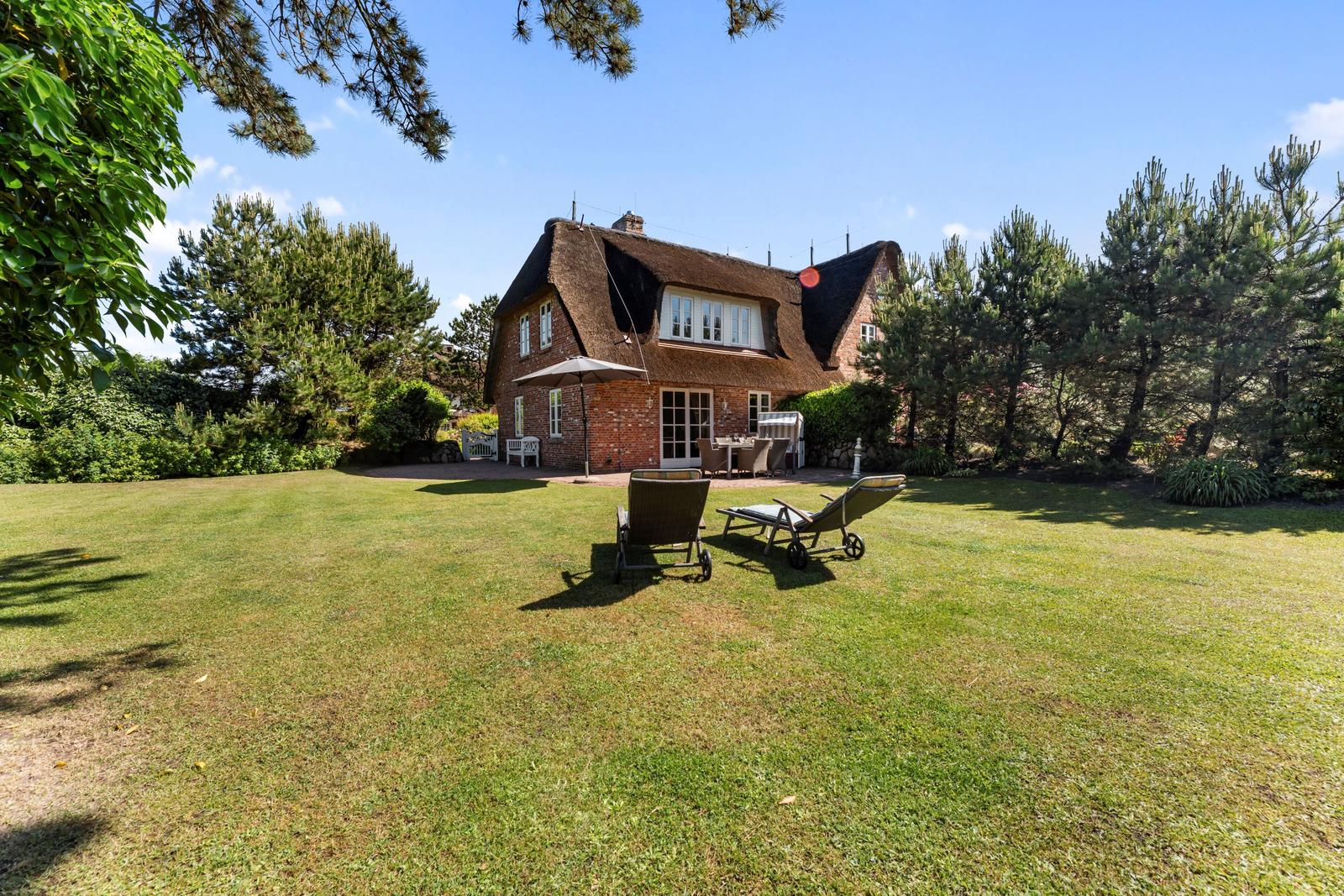 Roter Backsteinbau mit Strohdach, Garten mit Liegestühlen und Bäumen.