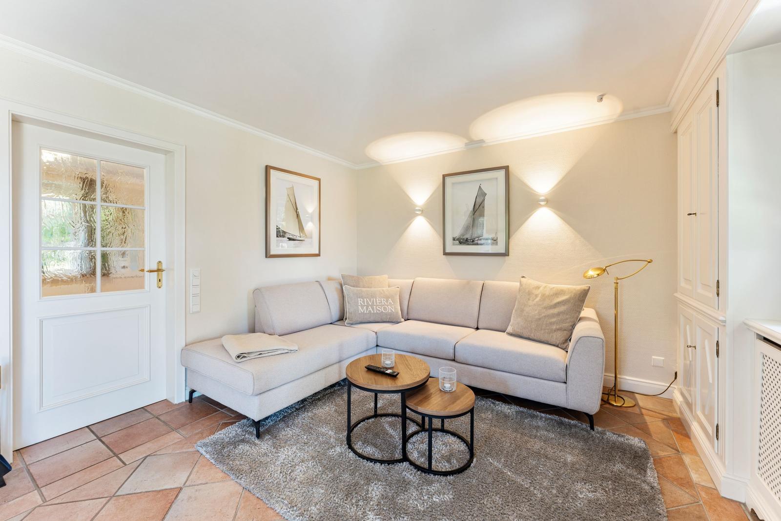 Wohnzimmer mit Ecksofa, zwei Tischen und Wandbildern. Tür und Schrank sind sichtbar.