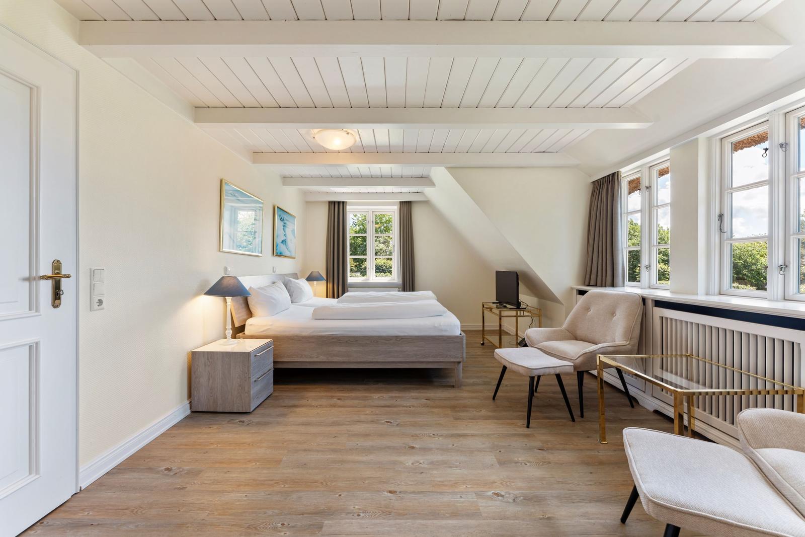 Schlafzimmer mit Doppelbett, Sitzgelegenheiten und großen Fenstern. Holzboden und weiße Wände.