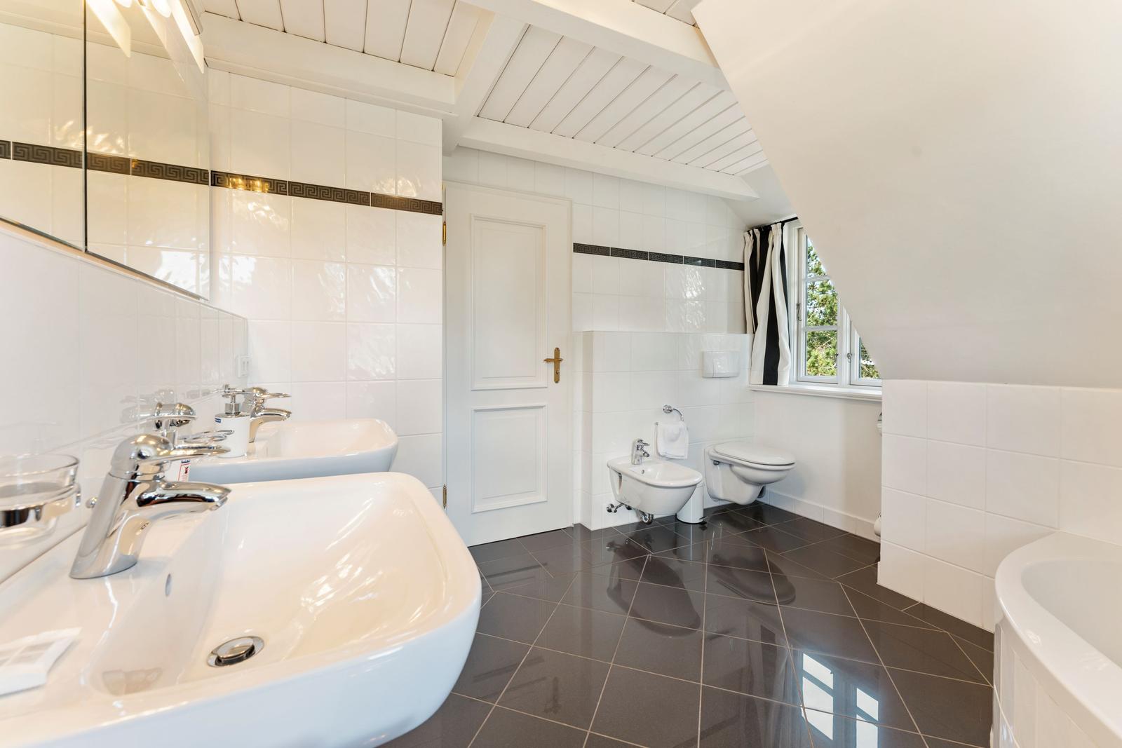 Badezimmer mit zwei Waschbecken, Toilette und Dusche. Weiße Wände und dunkler Boden.