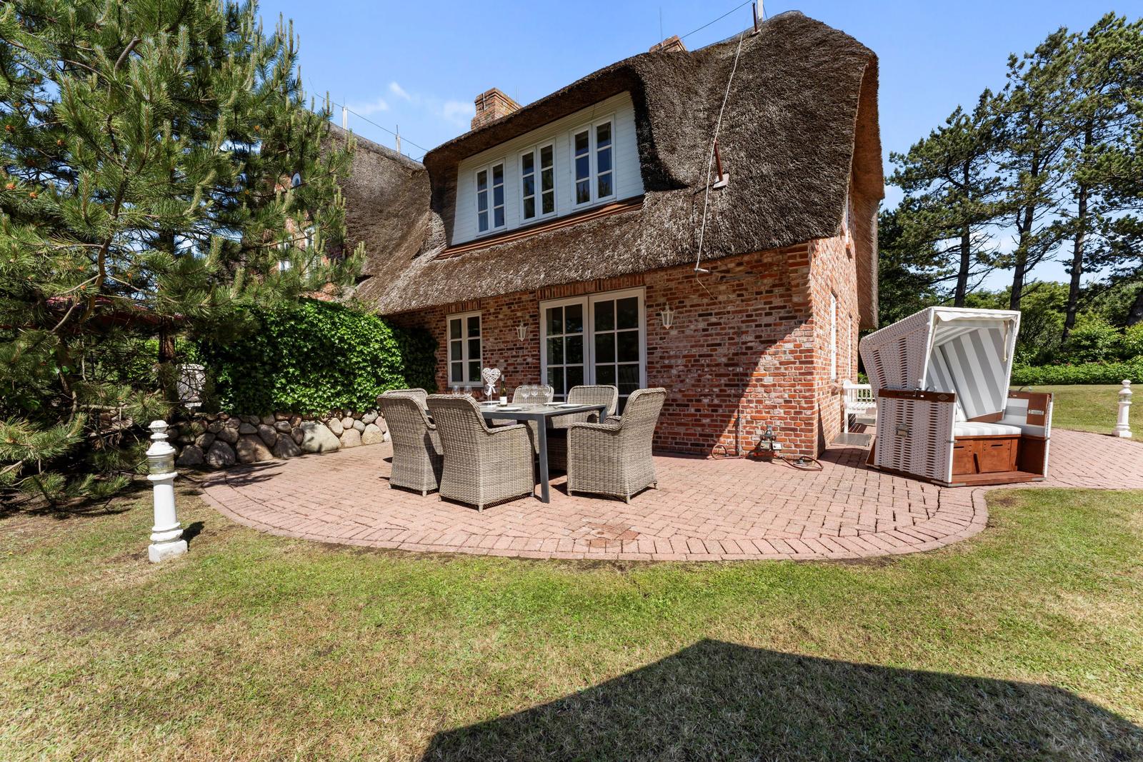 Haus mit Strohdach, Terrasse mit Tisch und Stühlen, Strandkorb und Grünflächen.