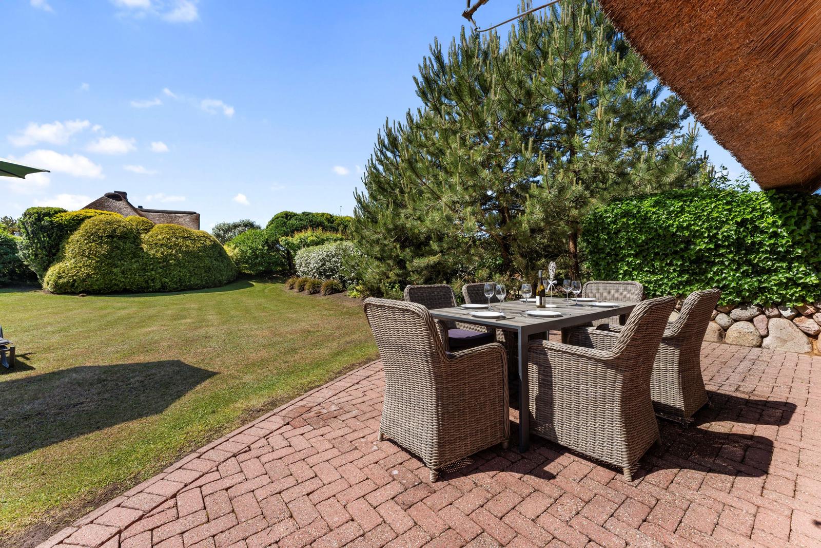 Terrasse mit Esstisch und Stühlen, umgeben von Grün und einem großen Baum.