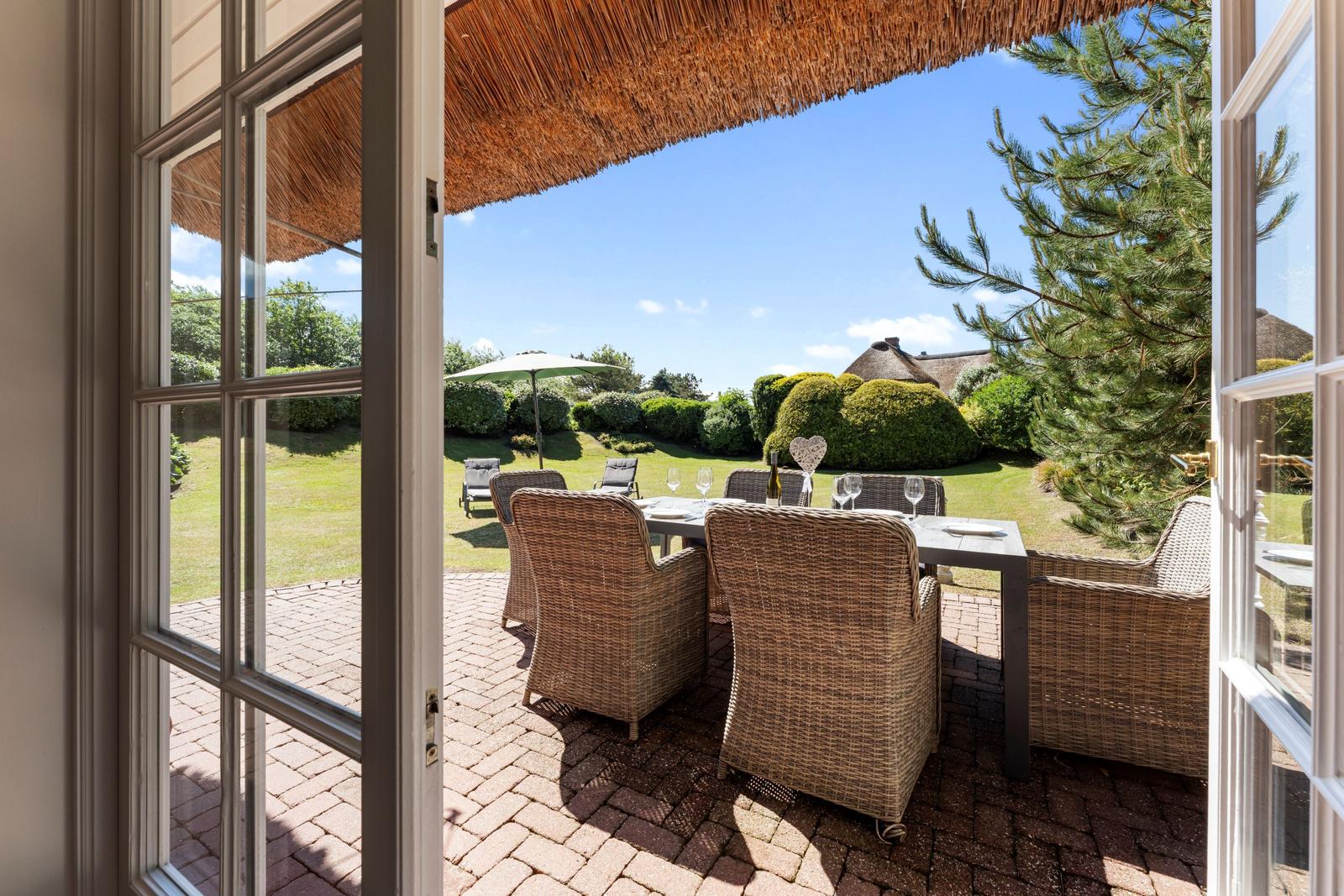 Terrasse mit Esstisch und Stühlen unter strohgedecktem Dach. Blick auf grünen Garten und Himmel.