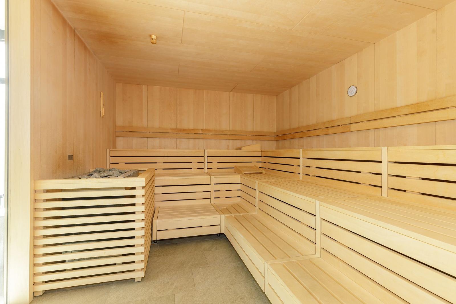 Sauna mit Holzbanken und Ofen, Wanduhr und Deckenleuchte sichtbar.
