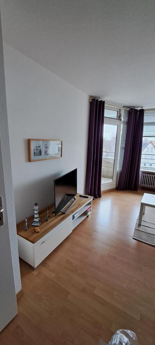 Wohnzimmer mit Holzboden, TV-Möbel und Balkontür mit lila Vorhängen.