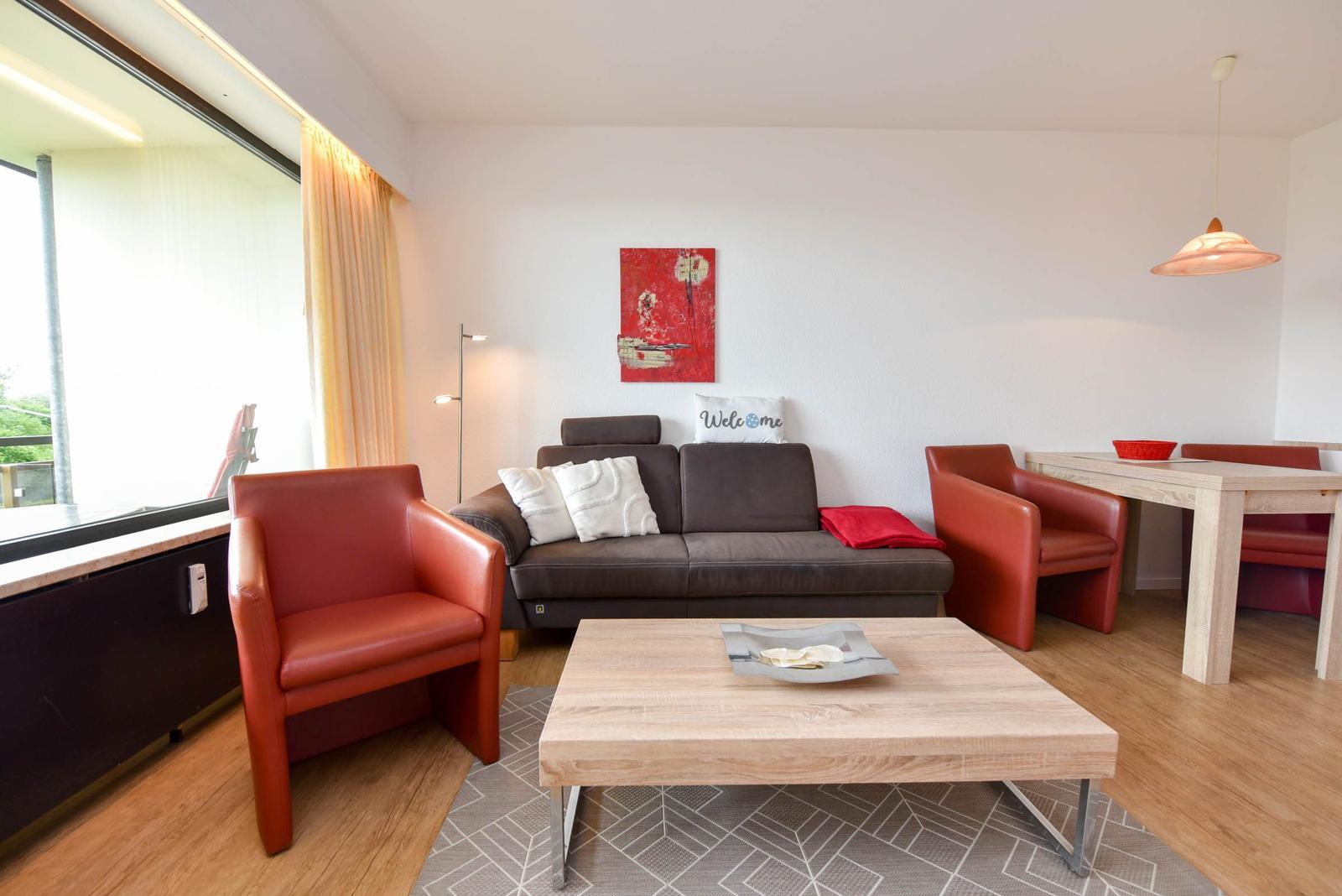 Wohnzimmer mit Sofa, zwei roten Sesseln und Esstisch. Große Fenster und Wandbild.