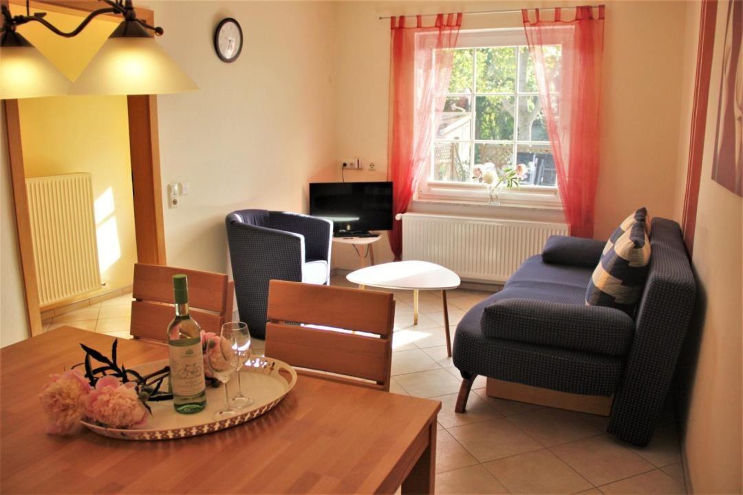 Living room with sofa, armchair, table, and TV. Window with red curtains. Table with wine and flowers.