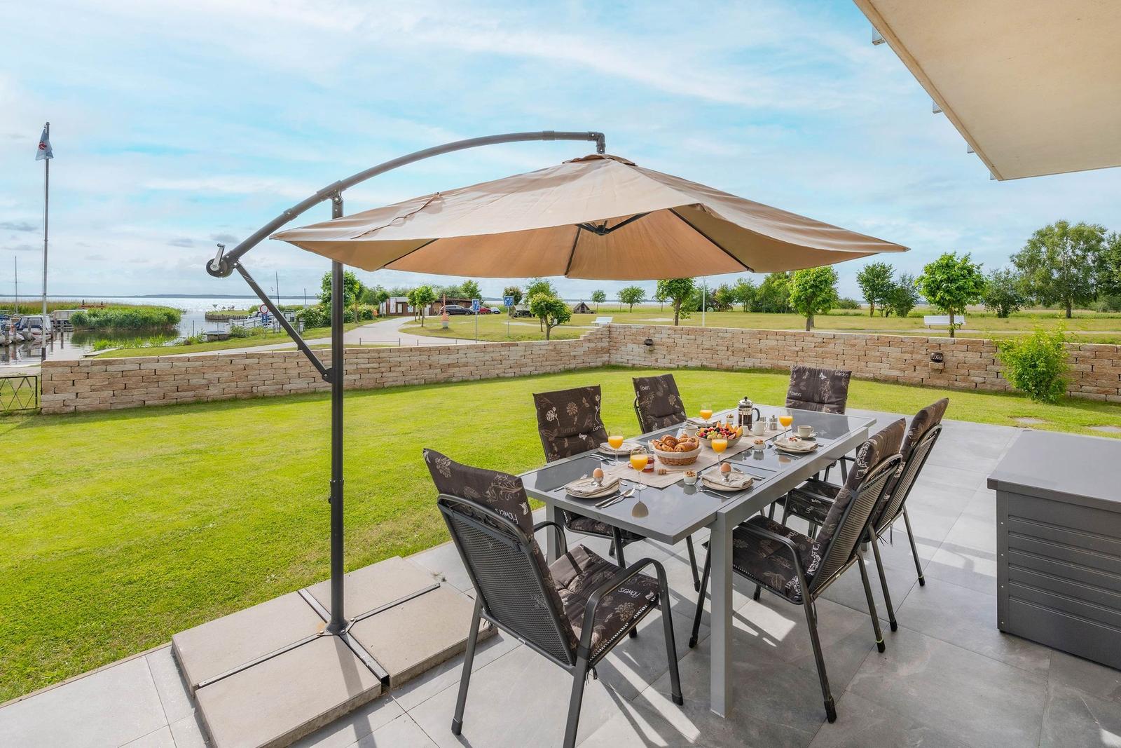 Terrasse mit Tisch, Stühlen und Sonnenschirm. Blick auf Grasfläche und Wasser.