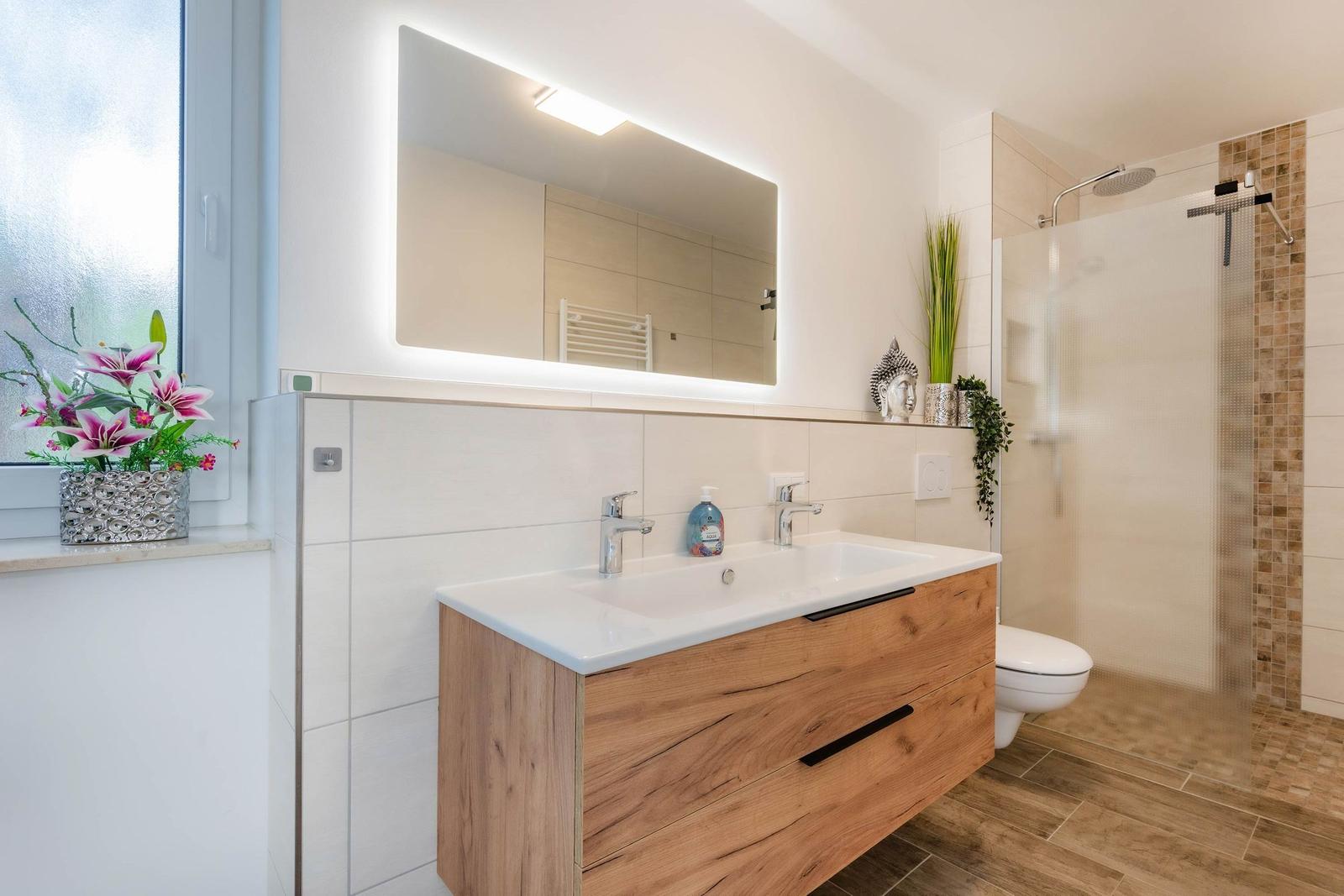 Badezimmer mit Waschtisch, Dusche und Fenster. Holzunterschrank und beleuchtetes Spiegel.