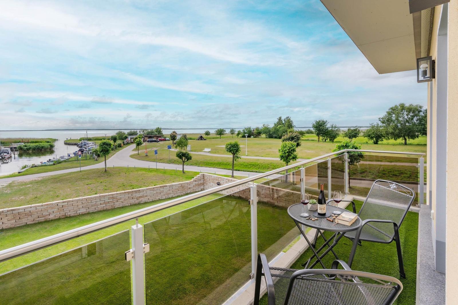 Balcony with table, chairs, and wine overlooking green park and water.