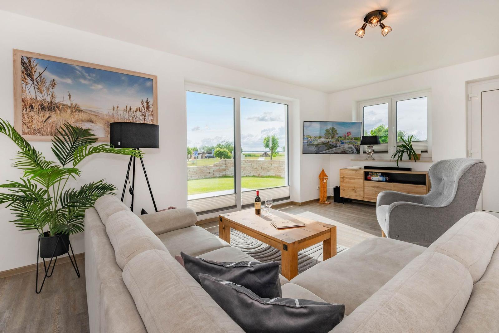 Wohnzimmer mit Sofa, Couchtisch und TV. Große Fenster mit Blick auf Grünfläche.