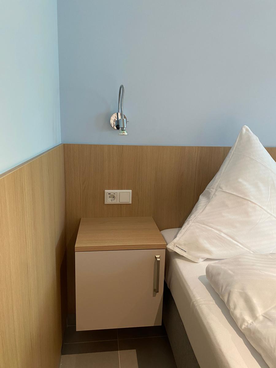 Bettkante mit Nachttisch und Wandlampe. Holzverkleidung und weiße Bettwäsche.