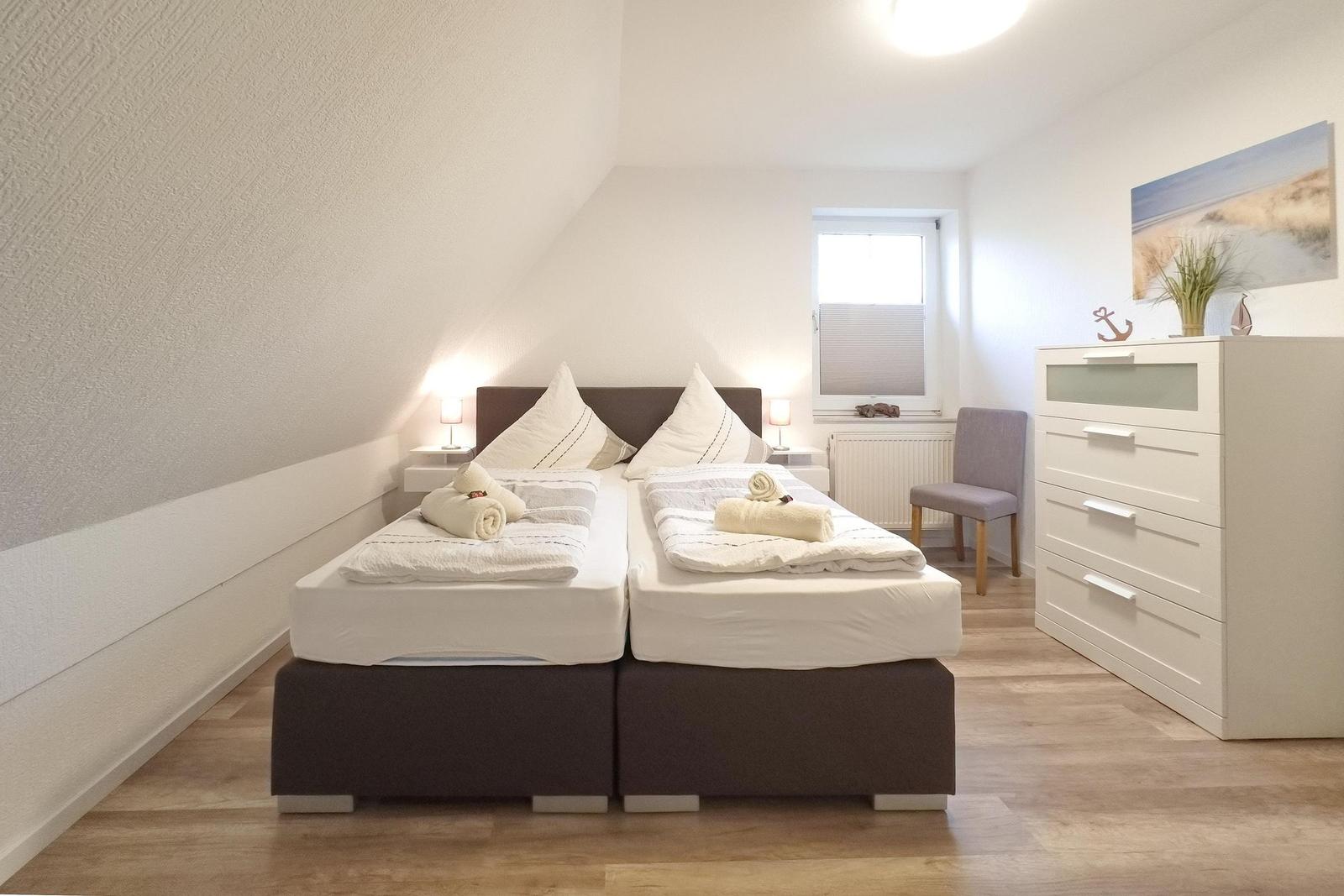 Zimmer mit Doppelbett, Schrank und Fenster. Holzboden und weiße Wände.