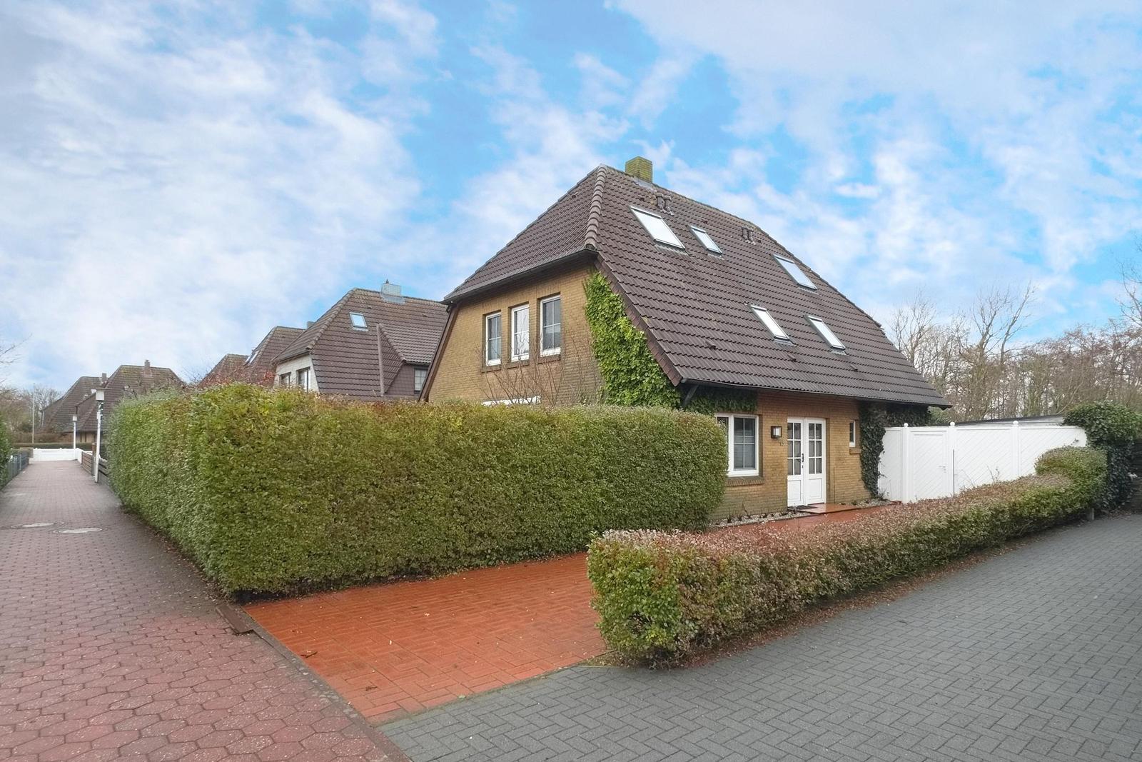Haus mit braunem Dach und grüner Hecke vor einem weißen Zaun.