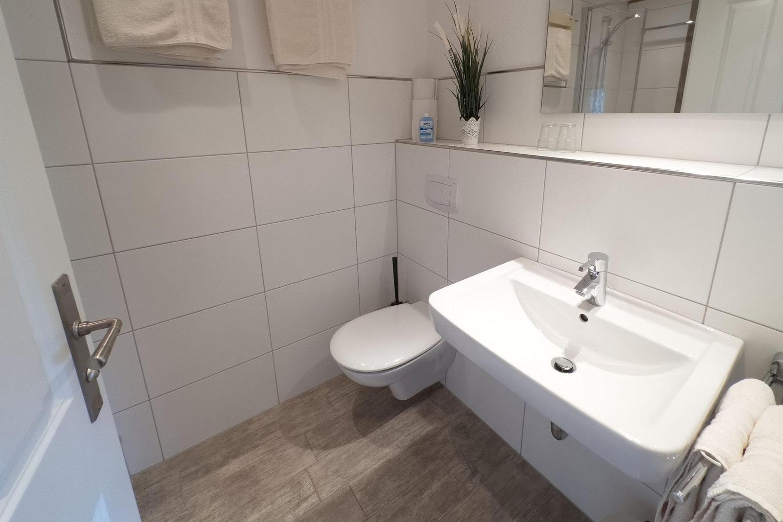 Badezimmer mit Toilette, Waschbecken und Spiegel. Holzoptik Boden. Handtücher und Pflanze.