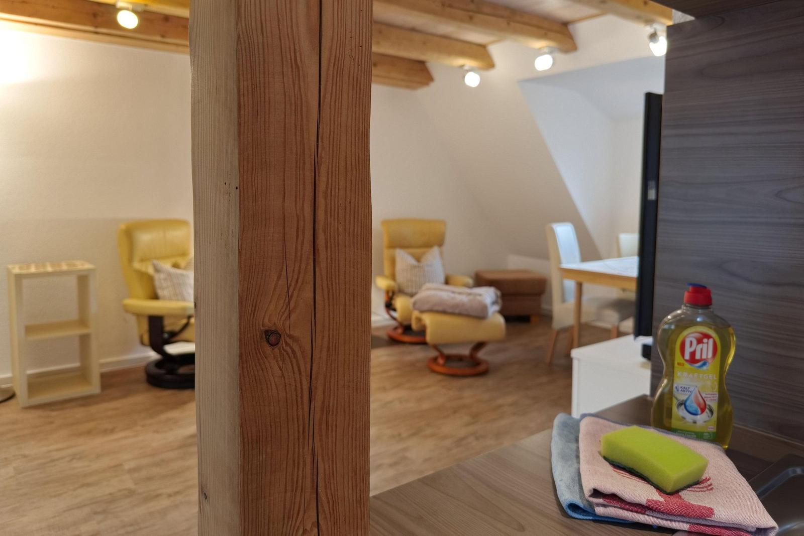Raum mit Holzbalken, gelben Sesseln und Küchentisch mit Spülmittel.