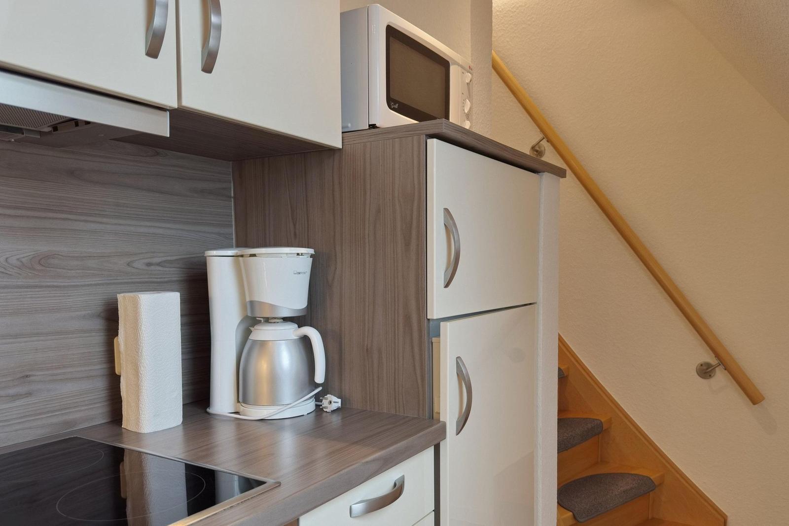 Küche mit Kaffeemaschine, Mikrowelle und Kühlschrank. Treppe mit Holzgeländer rechts.