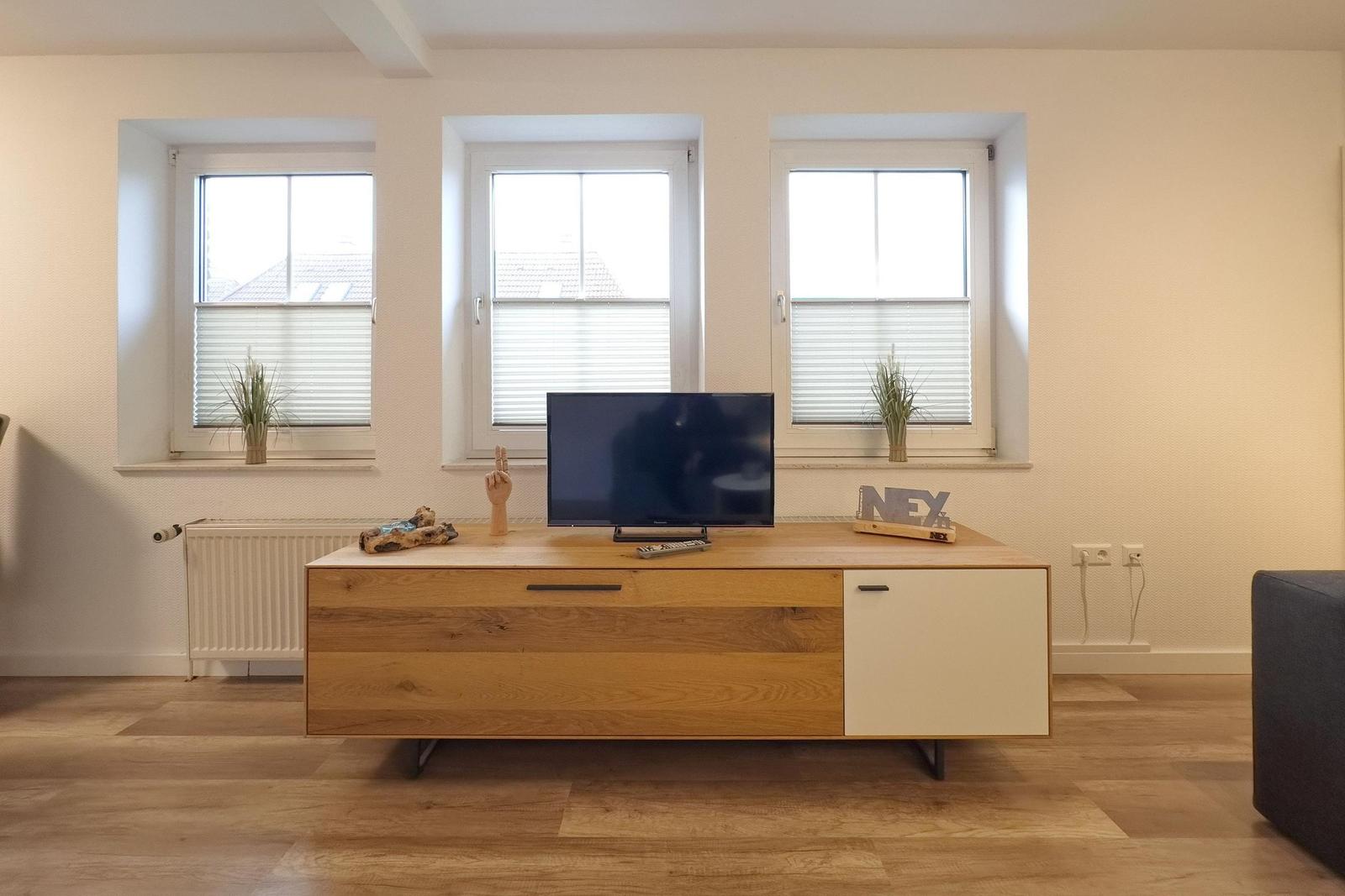 Wohnzimmer mit Holzregal, Fernseher und drei Fenstern mit Jalousien.